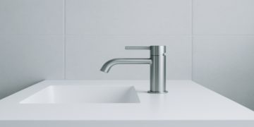 Sleek modern bathroom with a minimalist design. A metallic faucet extends over a white countertop with an inset sink, set against light gray tiled walls.