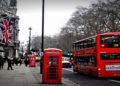 Street scene in London featuring a red double-decker bus and iconic phone booth. The British flag hangs on a building; trees line the overcast street.