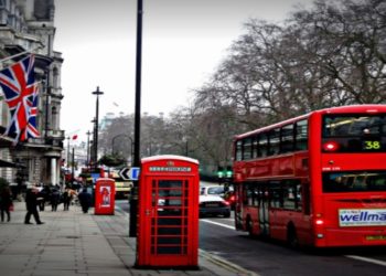 Street scene in London featuring a red double-decker bus and iconic phone booth. The British flag hangs on a building; trees line the overcast street.