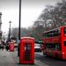 Street scene in London featuring a red double-decker bus and iconic phone booth. The British flag hangs on a building; trees line the overcast street.