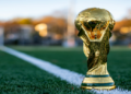 Golden trophy stands on a green soccer field near the white sideline, with a blurred background of trees in warm sunset light, evoking a sense of victory.