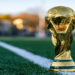 Golden trophy stands on a green soccer field near the white sideline, with a blurred background of trees in warm sunset light, evoking a sense of victory.