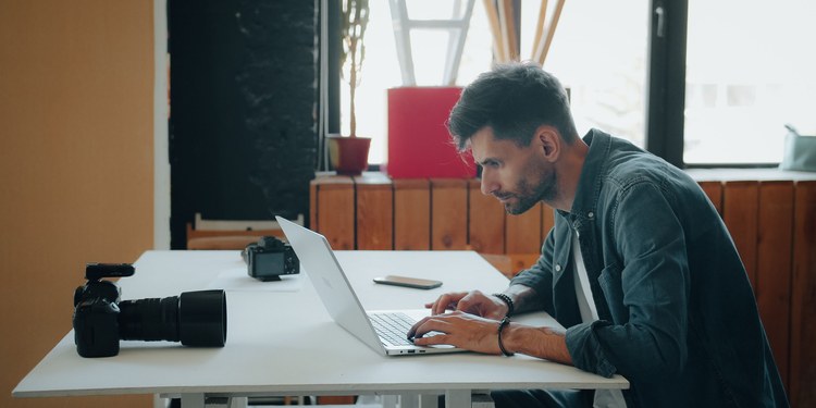 Photographer editing images on a laptop at a desk with cameras beside him
