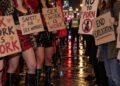 Protesters holding placards reading "Sex work is work" and "End exploitation" on a wet London street at night