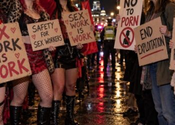 Protesters holding placards reading "Sex work is work" and "End exploitation" on a wet London street at night