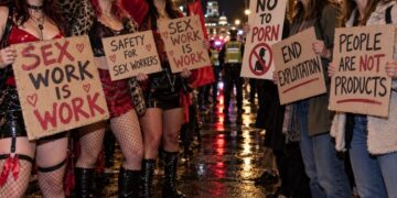 Protesters holding placards reading "Sex work is work" and "End exploitation" on a wet London street at night