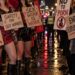 Protesters holding placards reading "Sex work is work" and "End exploitation" on a wet London street at night